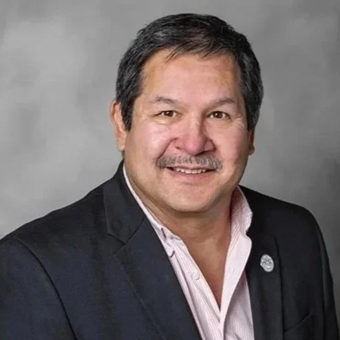 A middle-aged man with dark hair and a mustache, wearing a dark blazer and a light-colored shirt, smiling at the camera.