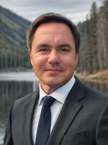 Headshot of a man in a suit and tie outdoors with a natural landscape of trees and mountains in the background.