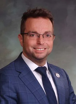 Headshot of a smiling man in a blue checkered suit, wearing glasses, with short brown hair, against a gray background.
