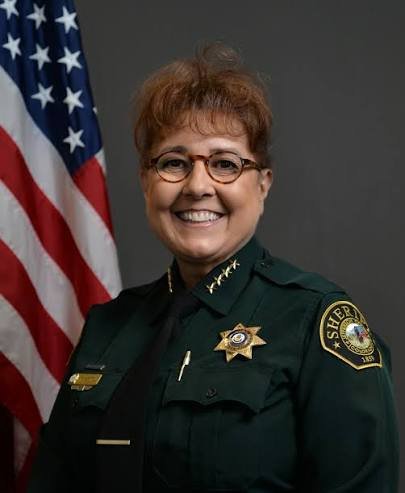 A female sheriff officer in uniform standing in front of an American flag, smiling at the camera.