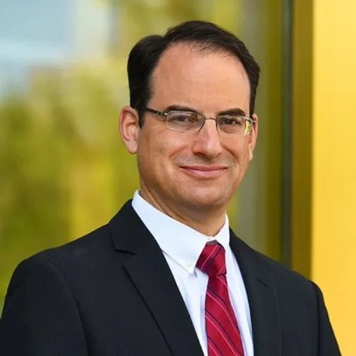 A man in a suit wearing glasses, standing outdoors with a yellow background.