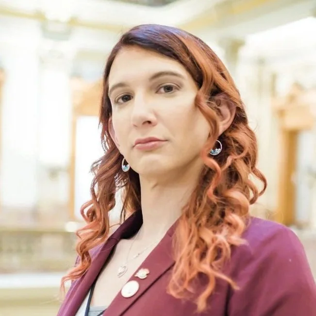 A woman with reddish-brown curly hair wearing a maroon blazer and earrings, standing indoors with a blurred background.