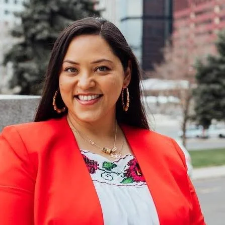 Woman with long dark hair smiling, wearing a red blazer and colorful embroidered blouse outdoors.