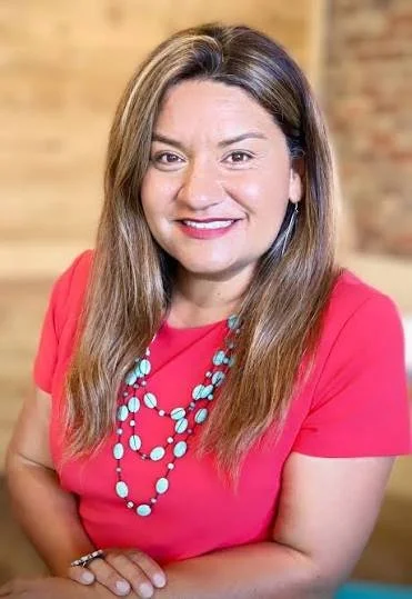 A woman with long, light brown hair wearing a red top and a turquoise beaded necklace, smiling at the camera, in an indoor setting with a blurred background.