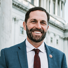 A smiling man in a blue suit and red tie standing outdoors in front of a government building.