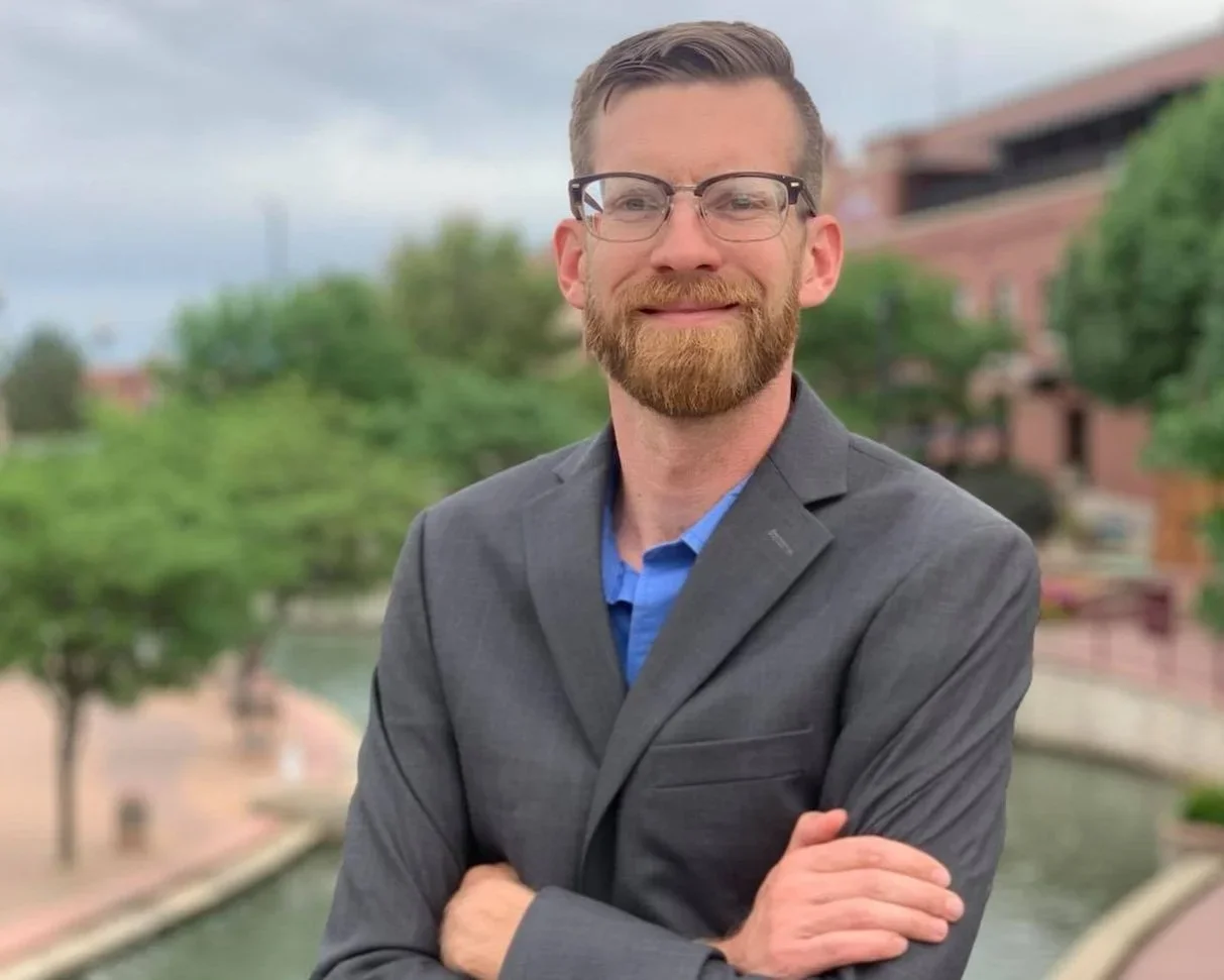 A man with glasses, a beard, and a mustache wearing a gray suit and blue shirt, standing outdoors with trees, a river, and buildings in the background.
