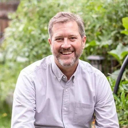 A smiling man with gray hair and a beard, wearing a light gray button-up shirt, outdoors surrounded by green foliage.