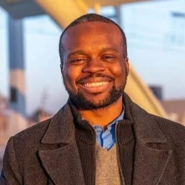 A smiling man with short hair and a beard, wearing a dark coat and a collared shirt, outdoors during daytime.