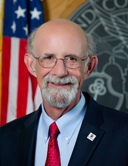 A man with glasses and a beard wearing a suit, red tie, and white shirt, standing in front of an American flag and a dark background with an emblem.