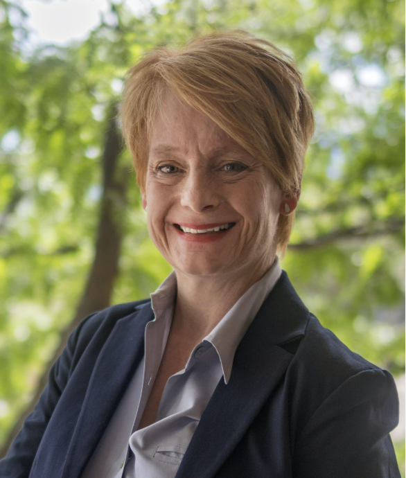 A woman with short, reddish hair, wearing a navy blazer and light blue shirt, smiling outdoors with a background of green trees.