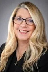 Professional woman with blonde hair, glasses, and black attire smiling against a gray background.