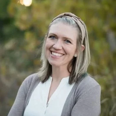 A woman with blonde hair smiling outdoors with blurred green trees in the background.
