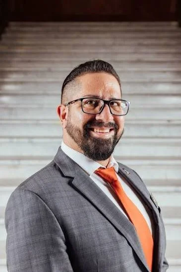 A man in a gray suit, white shirt, and orange tie standing on a staircase, smiling at the camera.