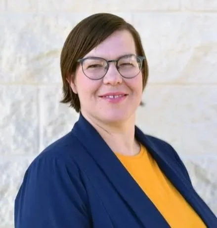 A woman with short brown hair, glasses, wearing a navy blazer and yellow top, smiling in front of a light-colored brick wall.