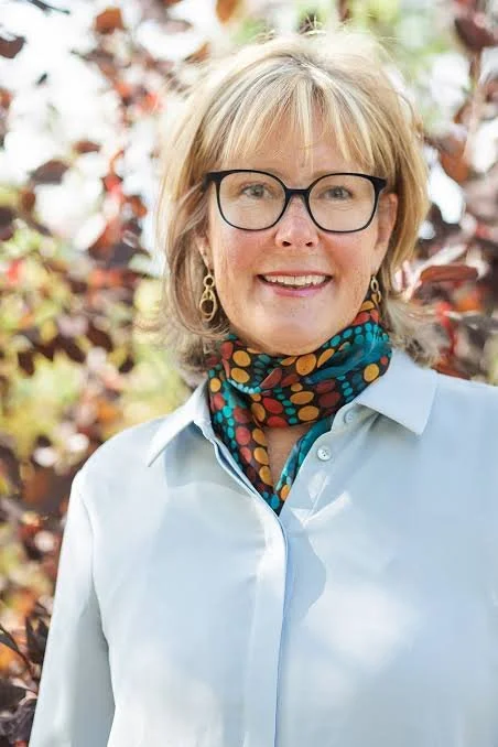 A woman with blonde hair, glasses, and earrings, smiling outdoors with blurred trees and leaves in the background, wearing a light blue shirt and a colorful polka dot scarf.
