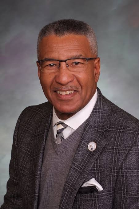 A smiling middle-aged man with short gray hair, glasses, wearing a suit jacket, a vest, a tie, and a white shirt, posing against a professional studio backdrop.