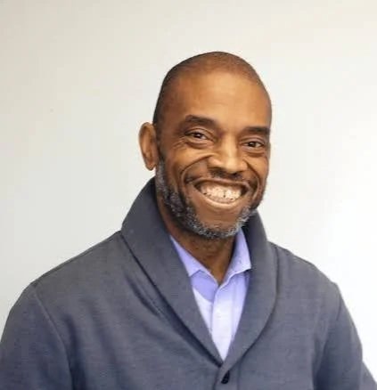 Smiling African American man with short hair and a beard, wearing a gray jacket and a light blue collared shirt, standing against a plain light-colored background.