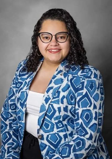 A woman with curly dark hair and glasses, wearing a blue patterned blazer and white top, smiling at the camera against a gray background.