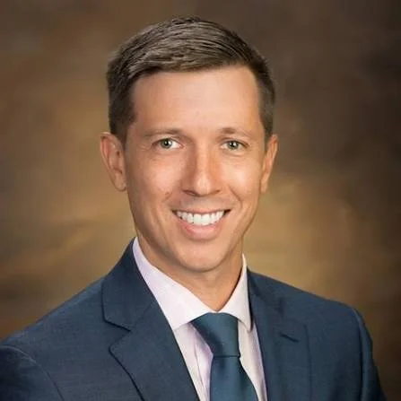 Headshot of a man in a blue suit, white shirt, and blue tie, smiling against a brown background.