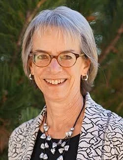 A smiling woman with short gray hair, glasses, wearing a patterned blazer and a necklace, outdoors with green foliage in the background.