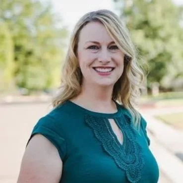 A smiling woman with shoulder-length blonde hair, wearing a teal blouse with lace details, standing outdoors with trees in the background.