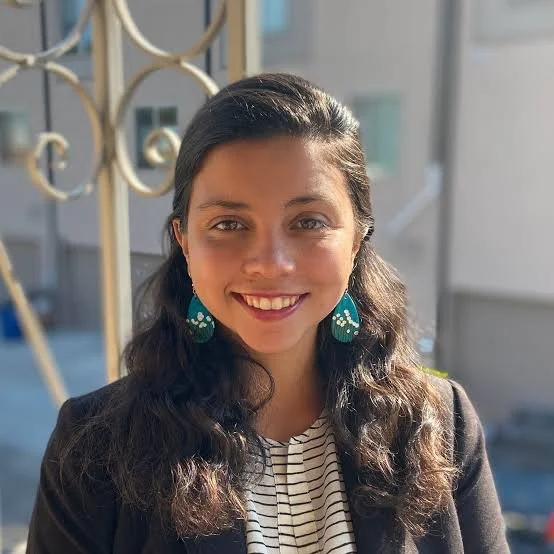 A young woman with dark hair, smiling, wearing teal earrings and a black blazer, standing outdoors with a decorative metal railing in the background.