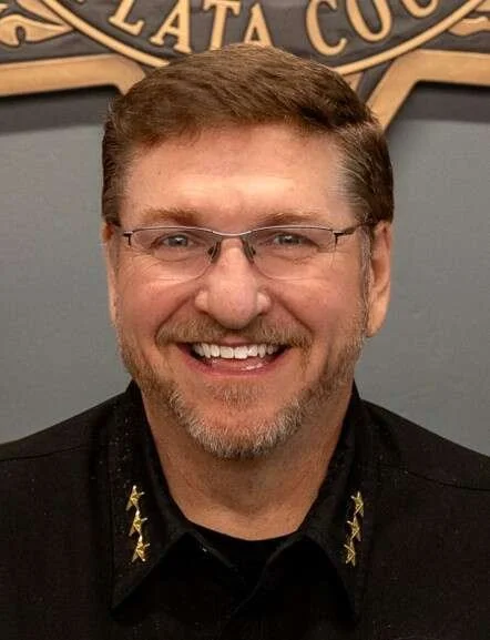 A man with glasses and a beard smiling in front of a badge or sign that says 'PLATA CO'.