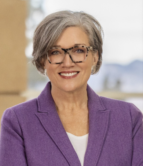 A smiling older woman with short gray hair, wearing glasses with a leopard print frame, a purple blazer, and a white shirt, standing outdoors with a blurred background.