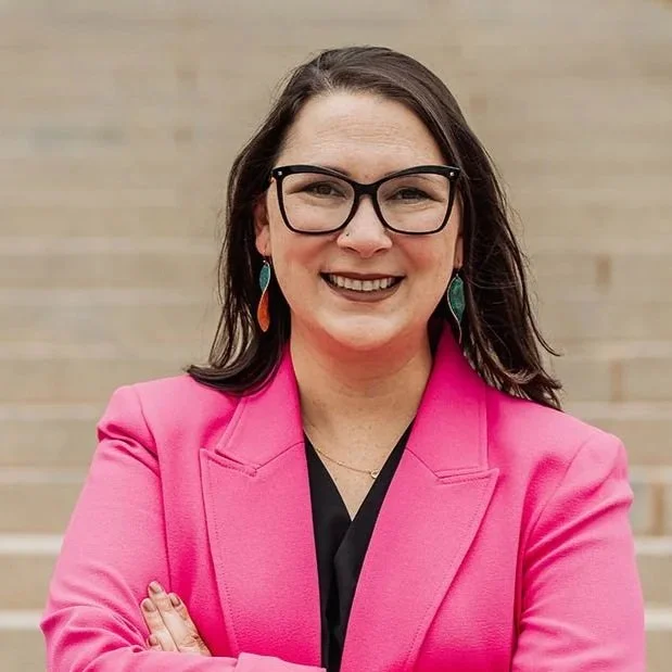 A woman with dark hair, glasses, and earrings wearing a bright pink blazer, smiling with arms crossed, standing against a neutral background.