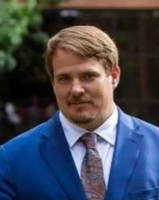 A man with light brown hair and a beard wearing a bright blue suit and a patterned tie, standing outdoors with a blurred background.