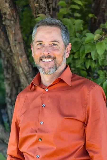 A smiling man with short gray hair and beard, wearing an orange button-up shirt, standing outdoors in front of trees and green foliage.