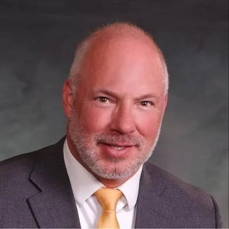 Professional headshot of a middle-aged man with light skin, a beard, and short gray hair, wearing a dark suit, white shirt, and a yellow tie.