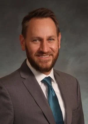 A man with short brown hair, a beard, wearing a gray suit, white shirt, and teal tie, smiling in a professional portrait.