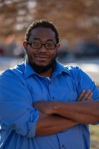 A man with glasses and a beard wearing a blue shirt, standing outdoors with his arms crossed and smiling.
