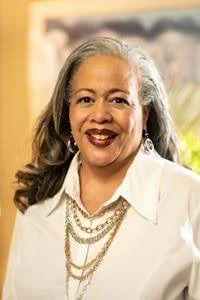 Portrait of a smiling woman with gray hair wearing a white blouse and layered necklaces.