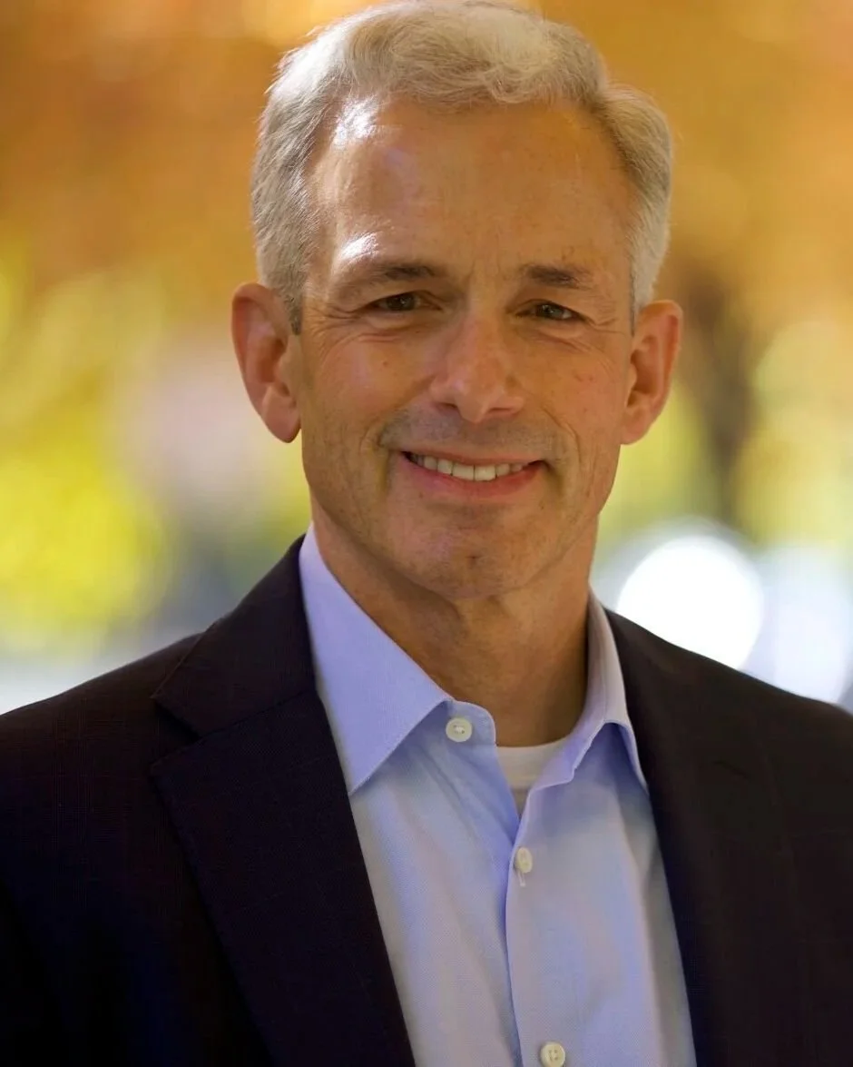 A middle-aged man with short gray hair, wearing a dark blazer and light blue dress shirt, smiling outdoors with blurred yellow and green foliage in the background.