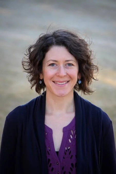 A woman with short, curly brown hair smiling outdoors, wearing a purple top and a black cardigan.