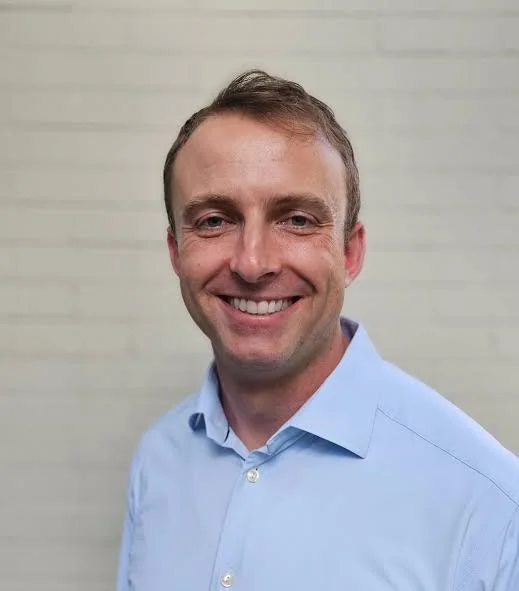 A smiling man with short light brown hair wearing a light blue collared shirt, standing against a light-colored wall.
