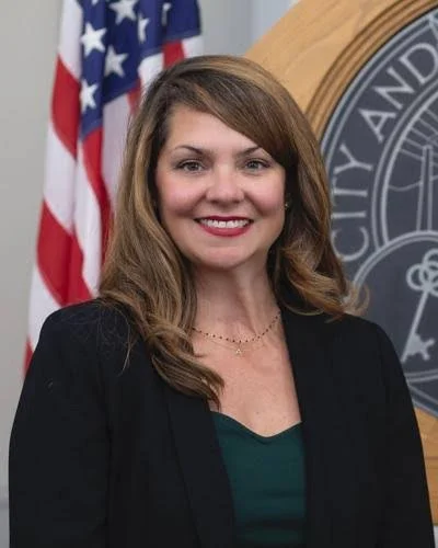 Woman with brown hair and a black blazer smiling, standing in front of an American flag and a city seal.