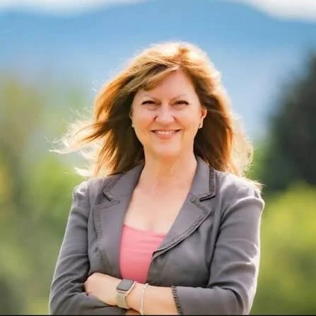 A woman with shoulder-length blonde hair smiling outdoors with mountains and trees in the background.