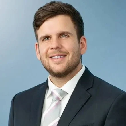 Portrait of a young man with short brown hair, wearing a dark suit, white shirt, and light-colored tie, smiling against a blue background.