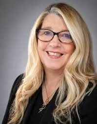 Professional woman with blonde hair, glasses, and black attire smiling against a gray background.