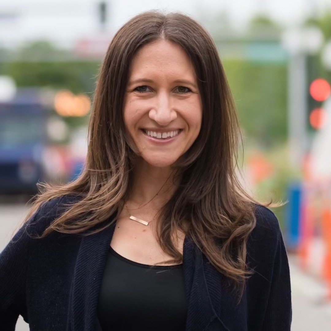 A woman with long brown hair smiling outdoors, blurred background with traffic lights and street scene.