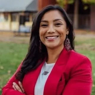 A woman smiling outdoors with her arms crossed, wearing a red blazer and earrings.