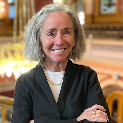 A smiling woman with curly gray hair standing indoors in a building with ornate woodwork and balconies.