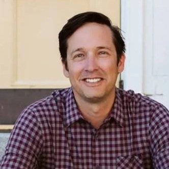 A man smiling, wearing a checkered shirt, sitting outside near a house.