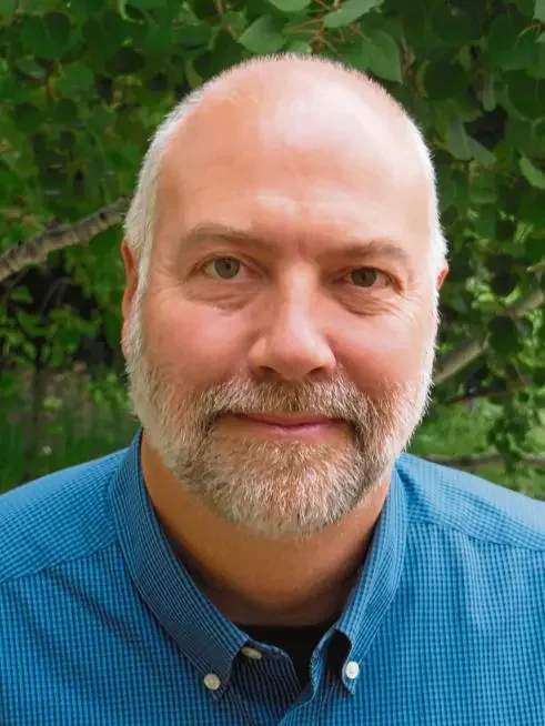 A middle-aged man with a beard and mustache, wearing a blue shirt, standing outdoors in front of green foliage.