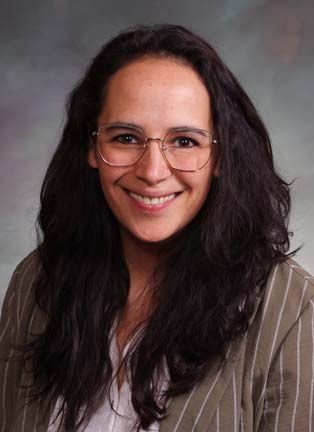 A woman with long, dark, curly hair and glasses, smiling at the camera, wearing a striped blazer.