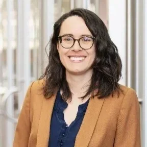 A woman with dark, curly hair wearing glasses, a blue blouse, and a tan blazer, smiling indoors.