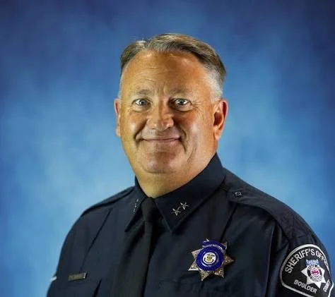 Portrait of a male police officer in uniform with a badge and shoulder patch, against a blue background.